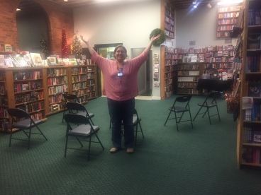 Picture of a bookstore a white woman with short brunette hair in a red shirt and jeans stand in the middle of a room with green carpet with her arms up and out. There are chairs behind her, and bookcases surround the outside of the room.