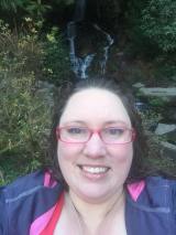 Selfie of a white fat woman with long brunette hair, pink glasses, and a grey and pink jacket in the foreground with a waterfall, rocks, and bushes in the background.