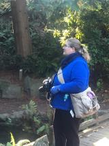 Picture with woods, rocks and greenery on the left and a fat blond woman in a blue sweater, gloves, and scarf and black pants holding a camera with a camera bag across her body.